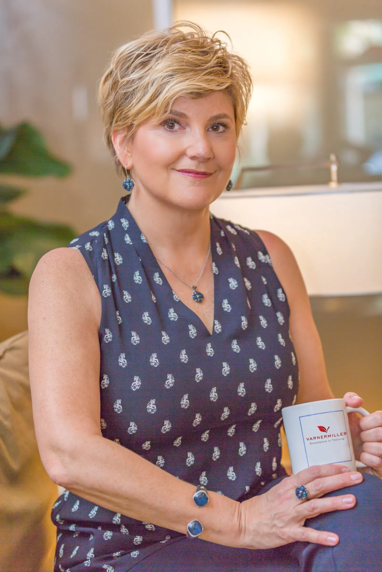 Lori Miller, owner, seated indoors holding a company-branded coffee mug and smiling at the camera.